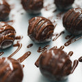 Close-up of 3 Banana Chocolate Protein Poppers on a tray, each drizzled with melted chocolate for a rich, glossy look. A decadently healthy snack packed with protein, shown with scattered chocolate drizzles.