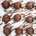 Three round Banana Chocolate Protein Poppers are arranged on a white surface, each drizzled with glossy melted chocolate in zigzag patterns—the perfect high protein, healthy snack for any time of day. More chocolate is artfully drizzled in the background.