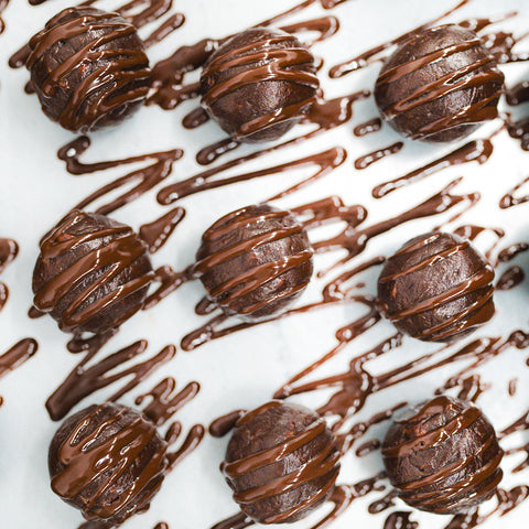 Three round Banana Chocolate Protein Poppers are arranged on a white surface, each drizzled with glossy melted chocolate in zigzag patterns—the perfect high protein, healthy snack for any time of day. More chocolate is artfully drizzled in the background.