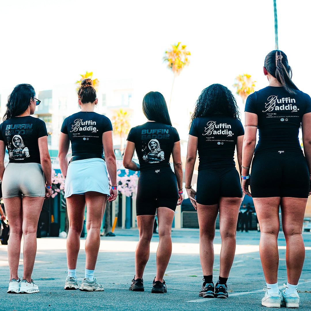 Five women stand outdoors with their backs to the camera, wearing black shirts reading Buffin Baddie. They stand on pavement with palm trees and buildings in the background under a blue sky.