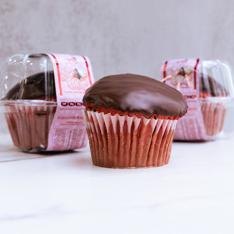 A Red Velvet Buffin Muffin sits in a red paper liner on a white surface, with two more high-protein Buffin Muffin VIP - Red Velvet muffins visible in clear plastic containers in the background.