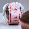 A clear plastic container labeled "Buffin Muffin Red Velvet" with a pink design and cupcake image holds a protein dessert. Branding is visible on the label, and a partially blurred red velvet muffin appears in the foreground.