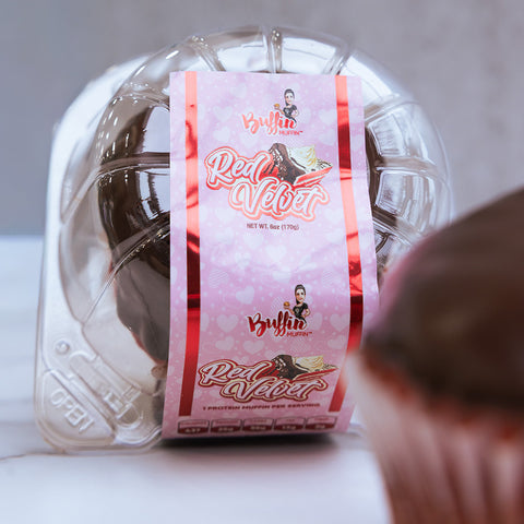 A clear plastic container labeled "Buffin Muffin Red Velvet" with a pink design and cupcake image holds a protein dessert. Branding is visible on the label, and a partially blurred red velvet muffin appears in the foreground.
