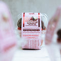 A clear plastic container holds a Buffin Muffin Red Velvet, topped with a pink label showing nutrition facts and the product name. The blurred pink and white background highlights this tasty high-protein dessert.