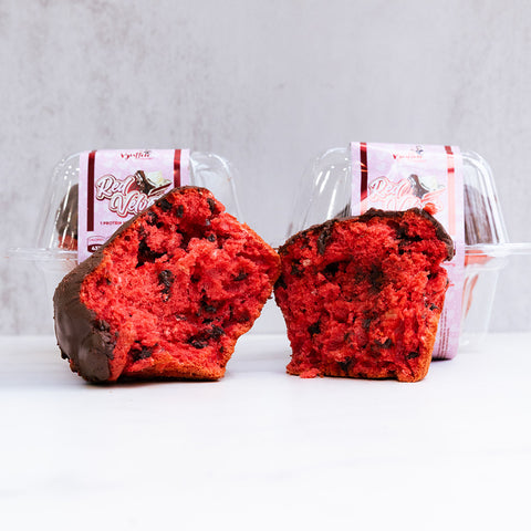 Buffin Muffin Red Velvet, sliced to show its moist inside and chocolate chips, sits before two clear containers labeled high-protein muffins on a plain white background.