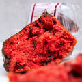 Close-up of a Red Velvet Buffin Muffin, vibrant and moist with chocolate chips, partially unwrapped from a clear plastic container. Chocolate pieces are scattered throughout this tempting protein dessert.