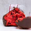 Buffin Muffin Red Velvet, a high-protein muffin with chocolate chips and a partial chocolate coating, rests on a white surface. A clear plastic container with label appears blurred in the background. Ideal for a tasty protein dessert.