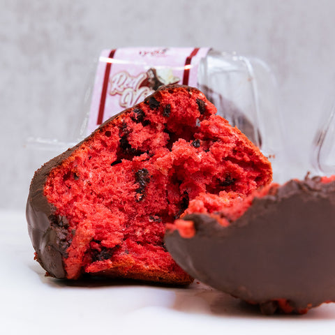 Buffin Muffin Red Velvet, a high-protein muffin with chocolate chips and a partial chocolate coating, rests on a white surface. A clear plastic container with label appears blurred in the background. Ideal for a tasty protein dessert.