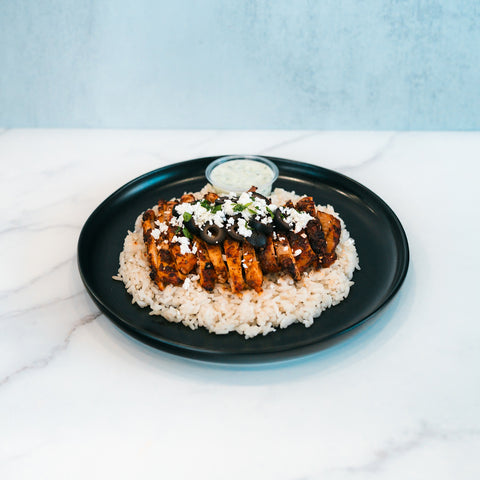 Olympian Chicken Gyro Bowl featuring sliced seasoned Hummus Fit chicken over rice, topped with crumbled cheese and sliced black olives, served with a side of sauce on a black plate.