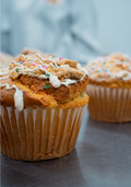 Close-up of a Dunk`d Out Exclusive-20 from Buffin Bakery, topped with white icing, colorful sprinkles, and crumbled cookie pieces, with another similar treat blurred in the background on a metal surface. Perfect for those seeking early-access products, this exclusive delight brings fun and flavor to any moment.