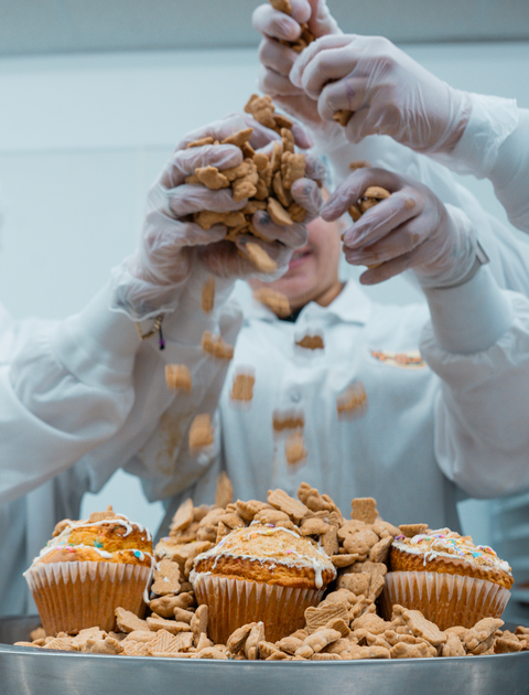 A group of people wearing white gloves carefully sprinkle Dunk`d Out Exclusive-20 animal-shaped crackers over muffins and onto a large pile in a metal tray, all within the bright, clean kitchen environment reminiscent of Buffin Bakery.