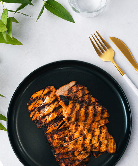 Grilled, sliced chicken breast with visible char marks served on a black plate, beside a gold fork and knife on a white tablecloth, with green leaves and a glass of water nearby.