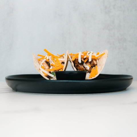 Baja Chicken Tacos feature grilled chicken, shredded cheese, and white sauce in low-carb tortillas, served upright on a black plate—a delicious high-protein meal prep option against a minimalist background.
