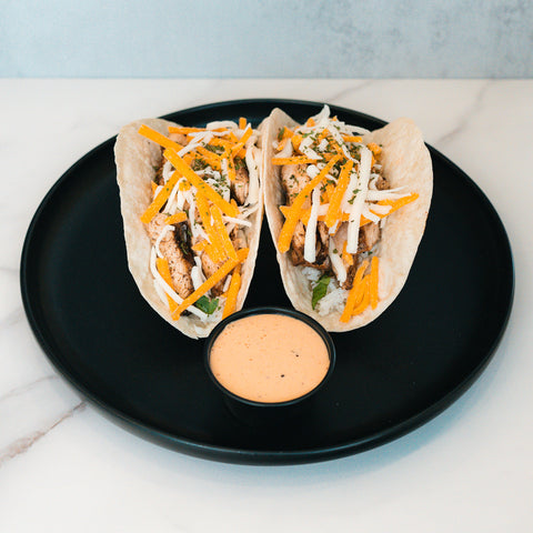Baja Chicken Tacos feature grilled chicken, cheddar and white cheese, and herbs in low-carb tortillas, served on a black plate with creamy dipping sauce—an ideal high-protein meal prep option.