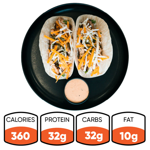 A black plate holds two Baja Chicken Tacos—shredded cheese and greens on low-carb tortillas with a dipping sauce. Nutrition: 360 calories, 32g protein, 32g carbs, 10g fat. Baja Chicken Tacos are ideal for high-protein meal prep.