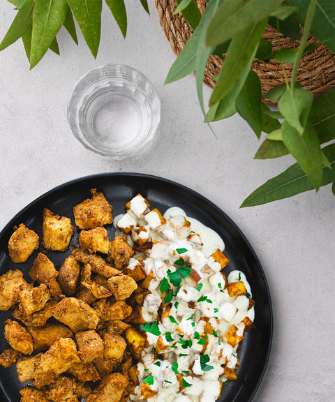 A black plate with seasoned grilled chicken pieces and roasted potatoes topped with a white sauce and herbs, next to a glass of water and green leafy plants on a light surface.
