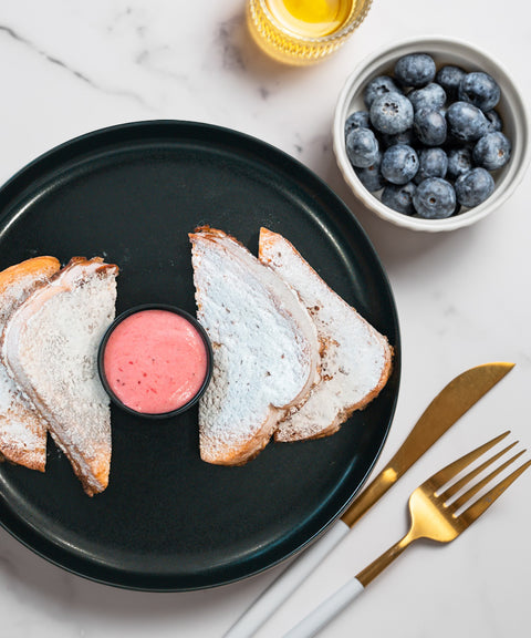 Three slices of powdered sugar-dusted toast with pink dipping sauce on a black plate, gold and white fork and knife, a bowl of blueberries, and a drink on a white marble surface.