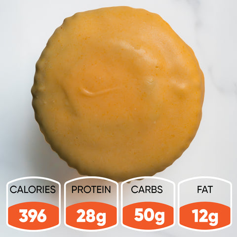 The Buffin Muffin VIP - Buffalicious Baddie, a round yellow-frosted high-protein muffin, sits on a white surface. Nutrition: 396 calories, 28g protein, 50g carbs, and 12g fat—an ideal gym-friendly snack.