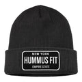The Hummus Fit Empire State Patch Beanie is a black, cuffed winter hat with a folded brim and a rectangular patch that reads "NEW YORK HUMMUS FIT EMPIRE STATE" in white—ideal for gym-to-street style or winter training.