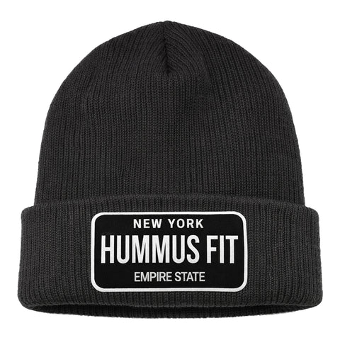 The Hummus Fit Empire State Patch Beanie is a black, cuffed winter hat with a folded brim and a rectangular patch that reads "NEW YORK HUMMUS FIT EMPIRE STATE" in white—ideal for gym-to-street style or winter training.