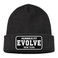 The Hummus Fit Evolve Cuffed Patch Beanie is a black rib-knit beanie with a folded brim and a rectangular patch reading HUMMUS FIT EVOLVE NEW YORK in bold white letters—ideal for athleisure or gym wear.