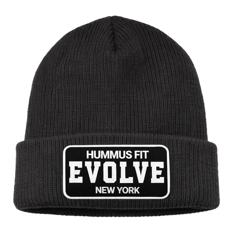 The Hummus Fit Evolve Cuffed Patch Beanie is a black rib-knit beanie with a folded brim and a rectangular patch reading HUMMUS FIT EVOLVE NEW YORK in bold white letters—ideal for athleisure or gym wear.