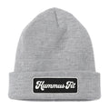 The Hummus Fit Vintage Patch Beanie is a light gray rib-knit style featuring a black rectangular patch on the front cuff with "Hummus Fit" in white cursive lettering.