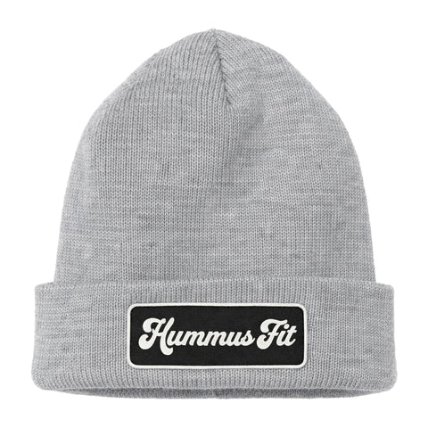 The Hummus Fit Vintage Patch Beanie is a light gray rib-knit style featuring a black rectangular patch on the front cuff with "Hummus Fit" in white cursive lettering.