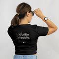 A woman with brown hair in a ponytail, wearing blue jeans and a smartwatch, stands facing away adjusting her sunglasses and rocking the Buffin Baddie Club Definition Crop Tee — Black.
