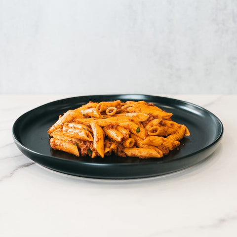 The Clean Bulk Pasta Bowl features penne pasta in red tomato sauce, topped with herbs. Ideal for healthy, high-protein meal prep, it’s perfect for pasta lovers. Served on a black plate over white marble with a neutral background.