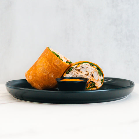 The Club Wrap, packed with chicken, greens, and veggies, is served halved on a black plate with dipping sauce—an ideal high-protein meal prep choice against a clean, light background.
