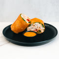 The Club Wrap, packed with protein-rich chicken, spinach, and red peppers, comes halved on a black plate with an orange dipping sauce on the side—ideal for meal prep or a quick lunch.