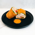 The Club Wrap, packed with high-protein chicken, spinach, and red bell peppers, is sliced in half and served on a black plate with orange dipping sauce—ideal for meal prep.