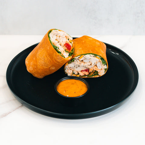 The Club Wrap, packed with high-protein chicken, spinach, and red bell peppers, is sliced in half and served on a black plate with orange dipping sauce—ideal for meal prep.