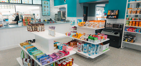 A bright, modern snack shop with teal walls displays a variety of packaged snacks on white shelves. In the background, staff work behind a counter with menus overhead. Shelves on the right hold more snacks and small appliances.