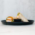 The Crispy Baked Chicken Wrap, halved and filled with lettuce, is a protein-rich lunch ideal for healthy meal prep. Served on a black plate with dipping sauce, it stands out against a simple light background.