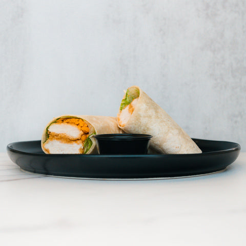 The Crispy Baked Chicken Wrap, halved and filled with lettuce, is a protein-rich lunch ideal for healthy meal prep. Served on a black plate with dipping sauce, it stands out against a simple light background.