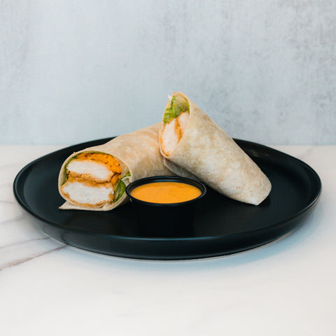 A sliced Crispy Baked Chicken Wrap filled with juicy breaded chicken, fresh lettuce, and sauce is served on a black plate with yellow dipping sauce—a high-protein lunch on a white marble surface.