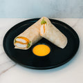 A Crispy Baked Chicken Wrap, filled with breaded chicken, lettuce, and cheese, is served halved on a black plate with yellow dipping sauce—an ideal high-protein lunch.