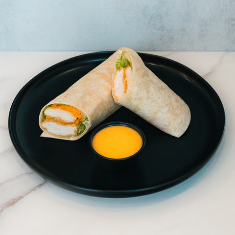 A Crispy Baked Chicken Wrap, filled with breaded chicken, lettuce, and cheese, is served halved on a black plate with yellow dipping sauce—an ideal high-protein lunch.