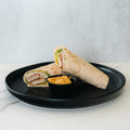 A black plate holds a healthy lunch: the Crispy Vegan Wrap, cut in half with lettuce inside, alongside a small black dish of orange dipping sauce on a white and gray surface.