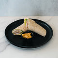The Crispy Vegan Wrap, filled with sliced plant-based chicken and lettuce, is served halved on a black plate with orange dipping sauce—a protein- and fiber-rich choice for a healthy lunch. The plate rests on white marble.