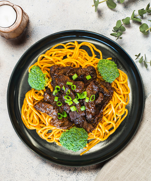 A black plate with yellow noodles topped with slices of glazed beef and chopped green onions, garnished with broccoli florets. The plate is set on a light surface with a candle and green plant nearby.