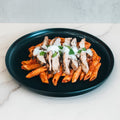 Fit A La Vodka With Chicken is served on a black plate, featuring penne pasta in red sauce, topped with grilled chicken slices, grated cheese, and green herbs on a white marble surface for a flavorful meal.