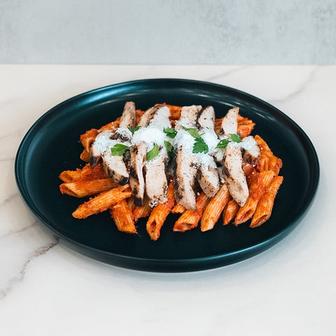 Fit A La Vodka With Chicken is served on a black plate, featuring penne pasta in red sauce, topped with grilled chicken slices, grated cheese, and green herbs on a white marble surface for a flavorful meal.