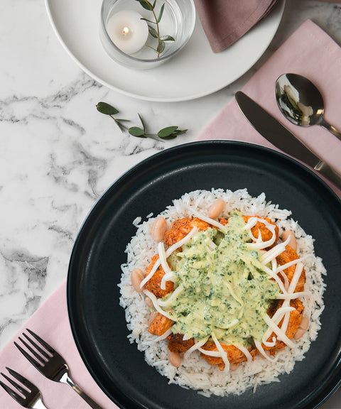 A black plate with white rice, seasoned chicken, white beans, shredded cheese, and green sauce, placed on a pink napkin with a fork and knife. A candle in a glass holder and a white plate are nearby on a marble surface.