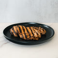 Competition Approved Grilled Chicken 1lb features halal, pre-cooked breast with char marks, sliced and arranged on a round black plate—ideal for quick meals or meal prep. Served atop a light marble surface for fresh presentation.