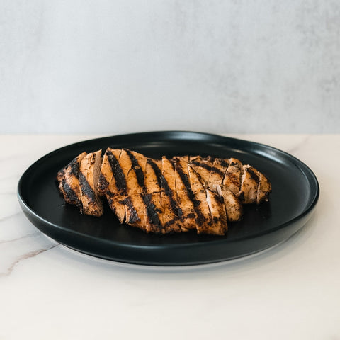 Competition Approved Grilled Chicken 1lb features halal, pre-cooked breast with char marks, sliced and arranged on a round black plate—ideal for quick meals or meal prep. Served atop a light marble surface for fresh presentation.