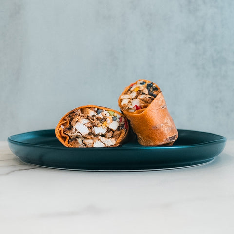 The Hey Arnold Burrito, a high-protein wrap packed with chicken, veggies, and cheese, is sliced in half and served on a dark blue plate against a light textured background—ideal for meal prep.