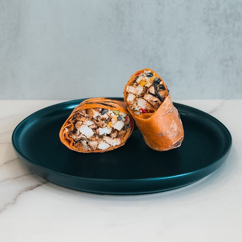 Two halves of the Hey Arnold Burrito—packed with chopped chicken, black beans, corn, and veggies—rest on a dark blue plate against a light backdrop. This high-protein meal is ideal for meal prep.